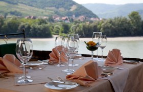 Gedeckter Tisch auf einer Restaurant-Terrasse mit Blick auf einen Fluss und bewaldete Hügel im Hintergrund.