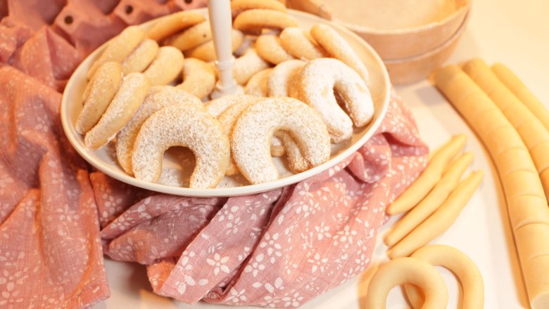 Ein Teller mit Vanillekipferln auf einem rosa Tuch, umgeben von Teigrollen.