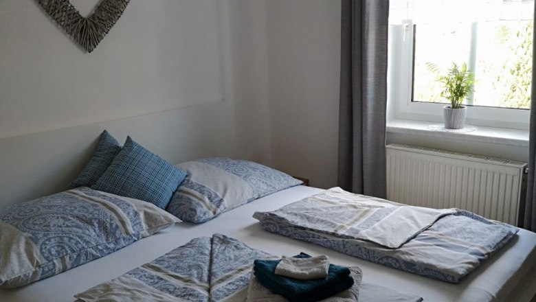 Ein gem&uuml;tliches Schlafzimmer mit einem Doppelbett, dekoriert mit blauer Bettw&auml;sche und Handt&uuml;chern. Ein Herzbild h&auml;ngt an der Wand, ein Fenster mit Vorhang daneben.