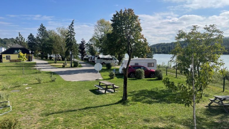 Ein Campingplatz mit Wohnwagen, Bäumen und einem See im Hintergrund bei sonnigem Wetter.