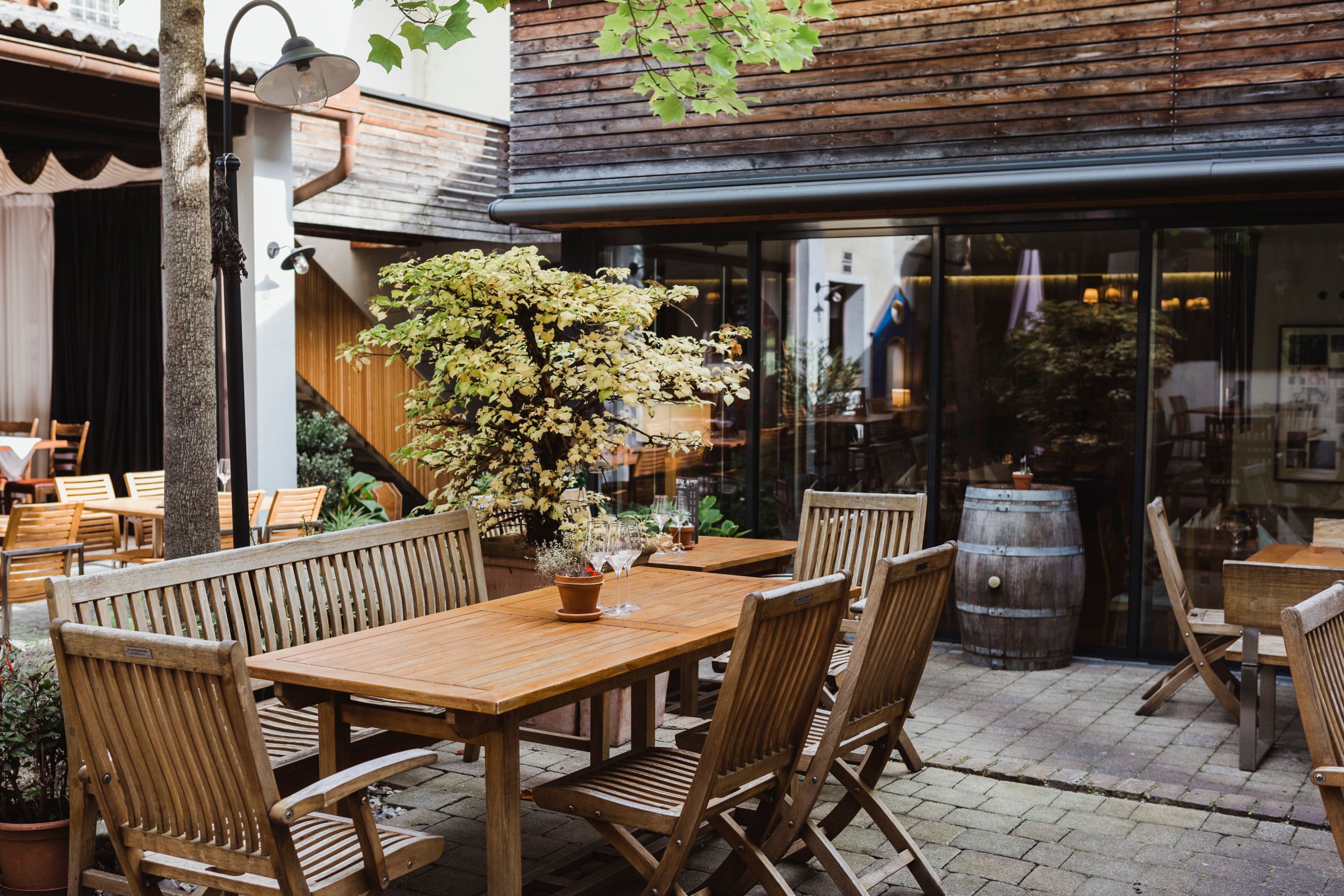 Ein gemütlicher Innenhof mit Holzmöbeln, Pflanzen und einem Weinfass, umgeben von Gebäuden mit Holzverkleidung.