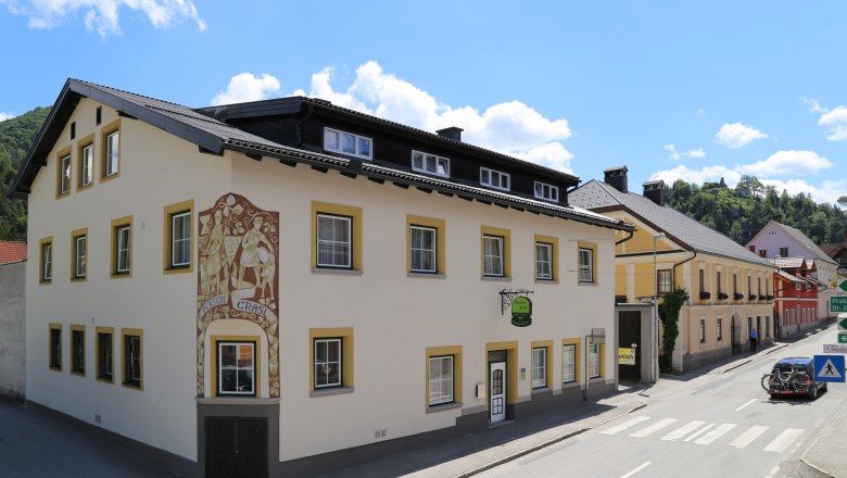 Ein gelbes Gebäude mit der Aufschrift 'Pension Grasl' an einer Straße bei sonnigem Wetter.