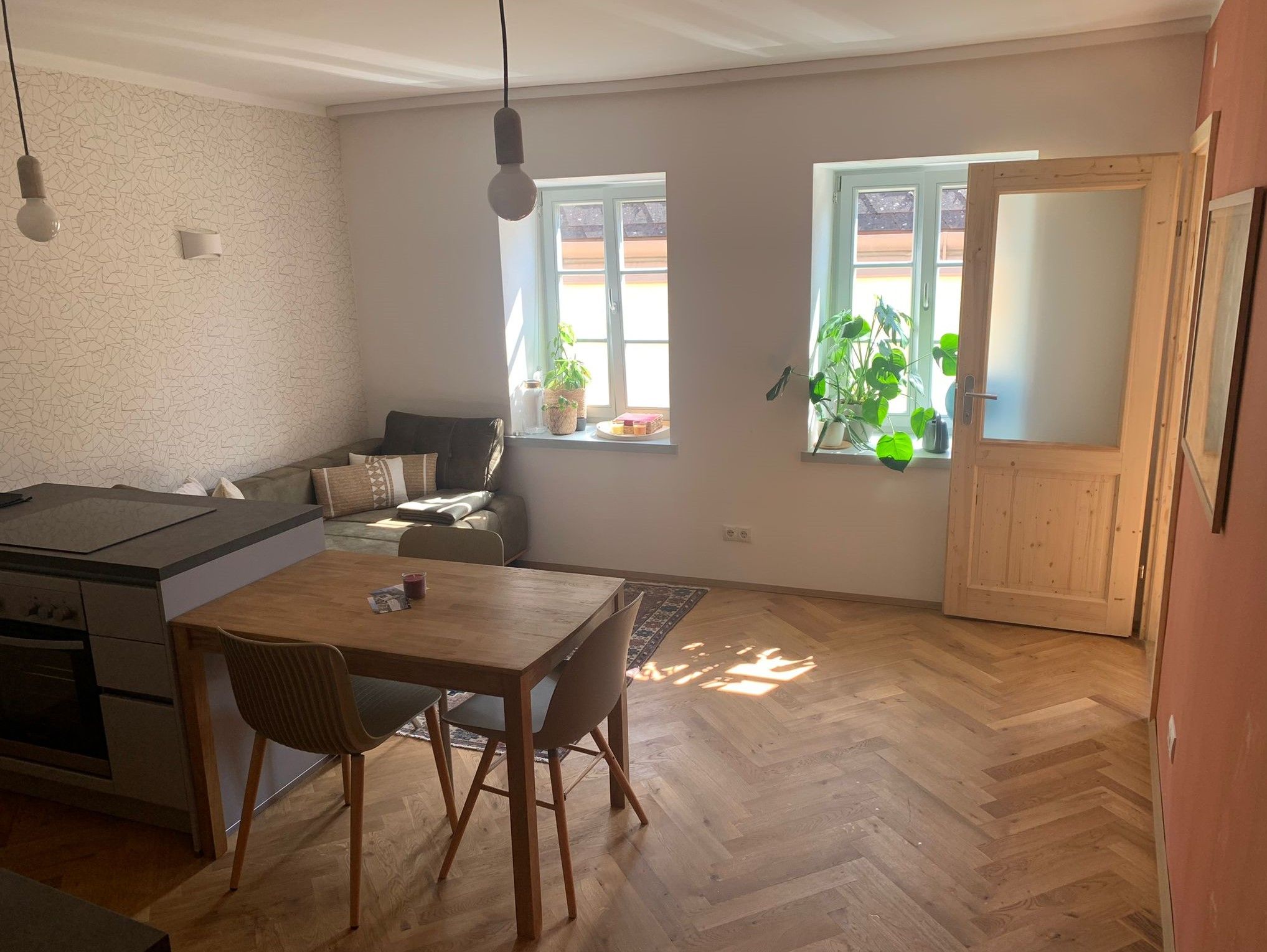 Helles Wohnzimmer mit Holzboden, Esstisch, Sofa und Pflanzen am Fenster.