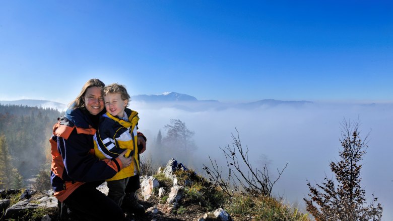 Eine Frau und ein Kind stehen auf einem H&uuml;gel mit Blick auf ein Nebelmeer und Berge im Hintergrund.