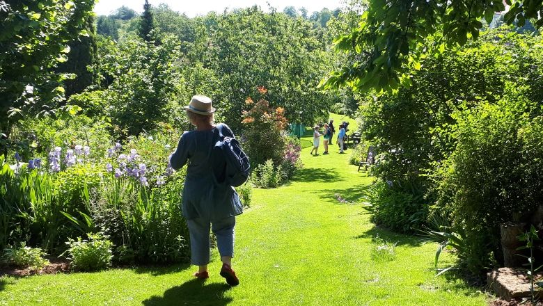 Person mit Hut spaziert durch einen grünen Garten mit Blumen und Bäumen.