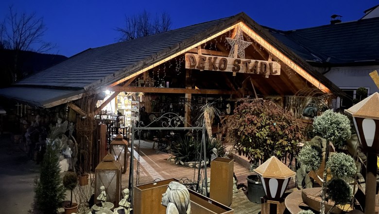 Ein dekorativ beleuchtetes Holzgebäude mit der Aufschrift 'Deko Stadl' bei Nacht, umgeben von Pflanzen und Dekorationsartikeln.