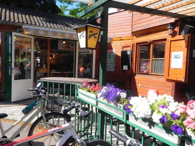 Ein gemütlicher Imbiss mit Holzwänden, Fahrrädern und blühenden Blumen im Vordergrund.