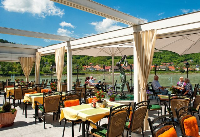 Restaurantterrasse mit Blick auf einen Fluss und gr&uuml;ne H&uuml;gel im Hintergrund.