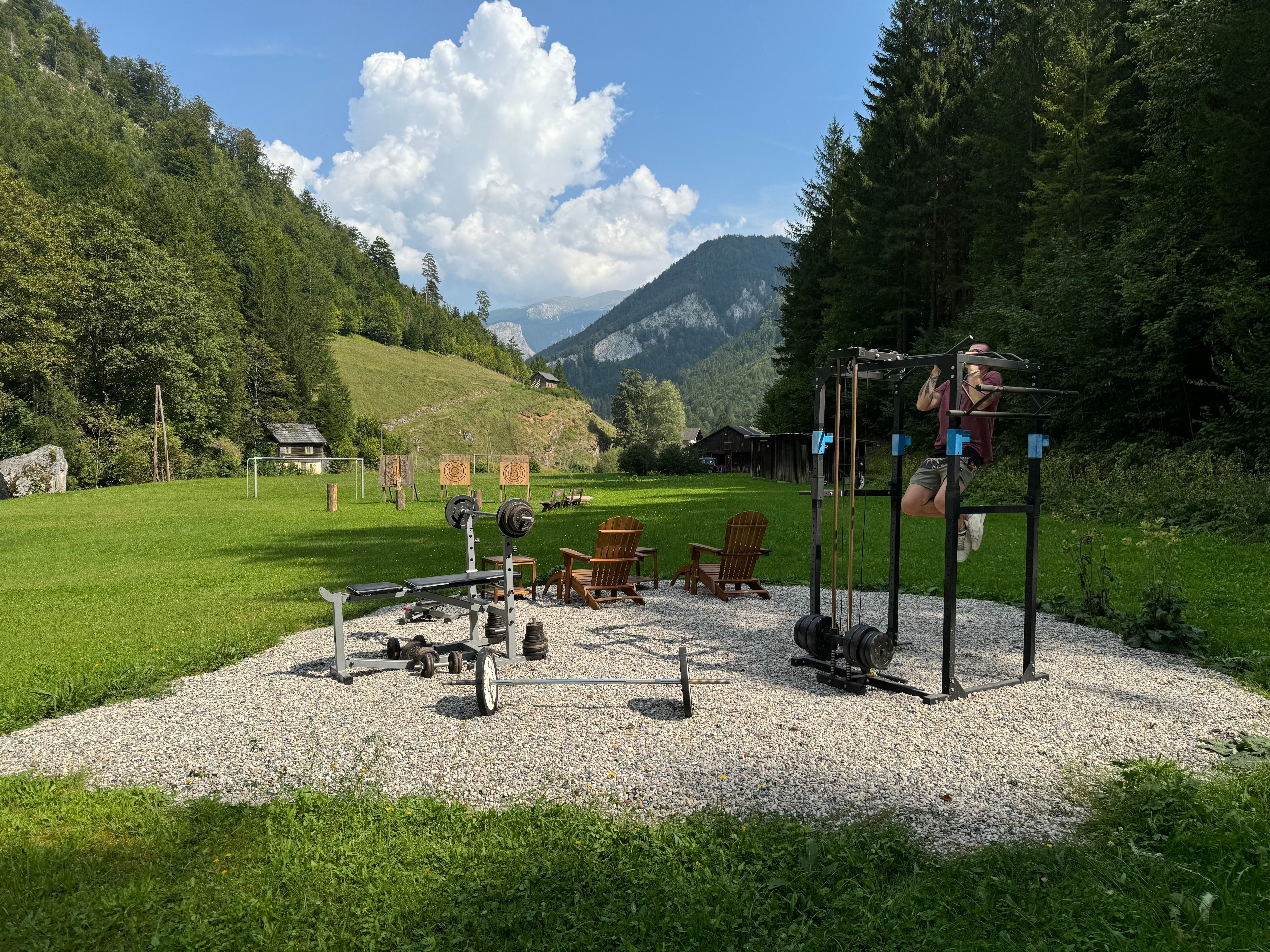 Outdoor-Fitnessbereich mit Trainingsgeräten in einer grünen, bergigen Landschaft.
