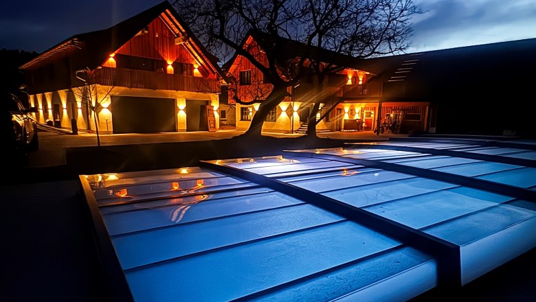 Nachtansicht eines beleuchteten Geb&auml;udes mit Baum und &uuml;berdachtem Bereich im Vordergrund.