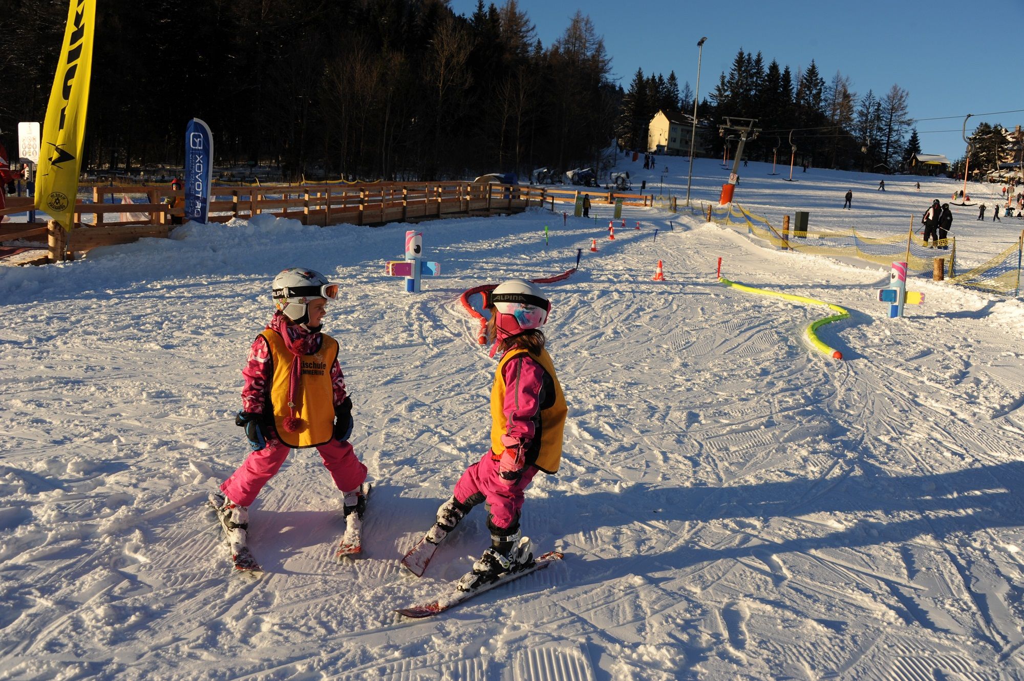 Zwei Kinder in Skiausrüstung im Schnee, umgeben von bunten Hindernissen.