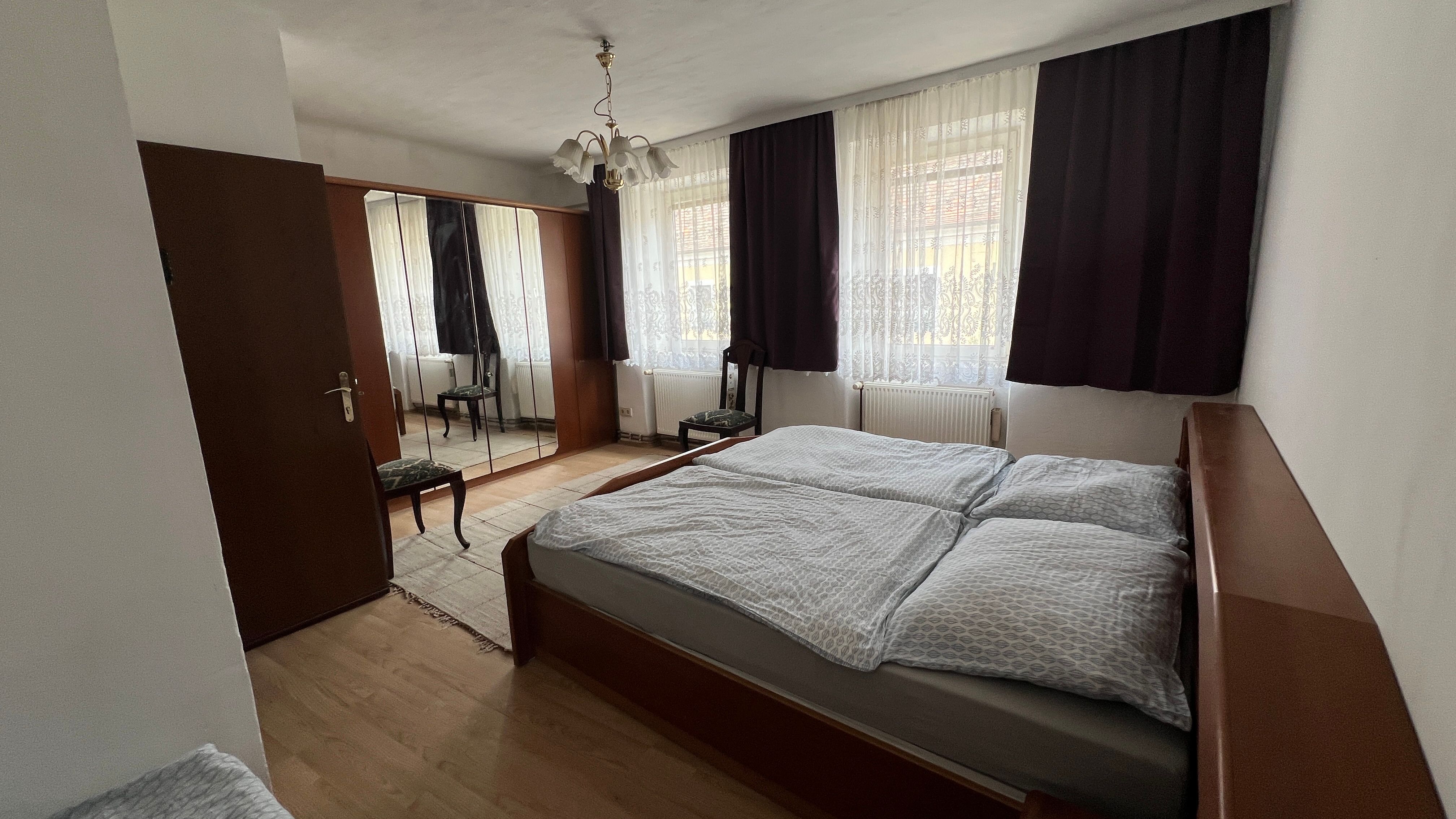 Schlafzimmer mit Doppelbett, Kleiderschrank mit Spiegel, Stuhl und Vorhängen an den Fenstern.