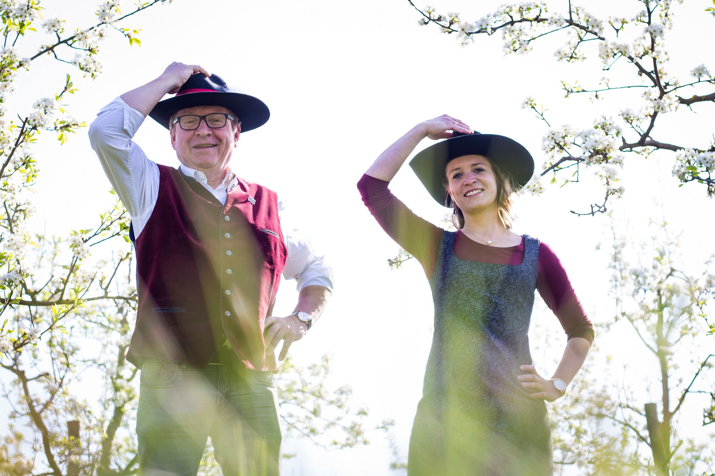 Zwei Personen in traditioneller Kleidung mit Hüten stehen in einem blühenden Obstgarten.