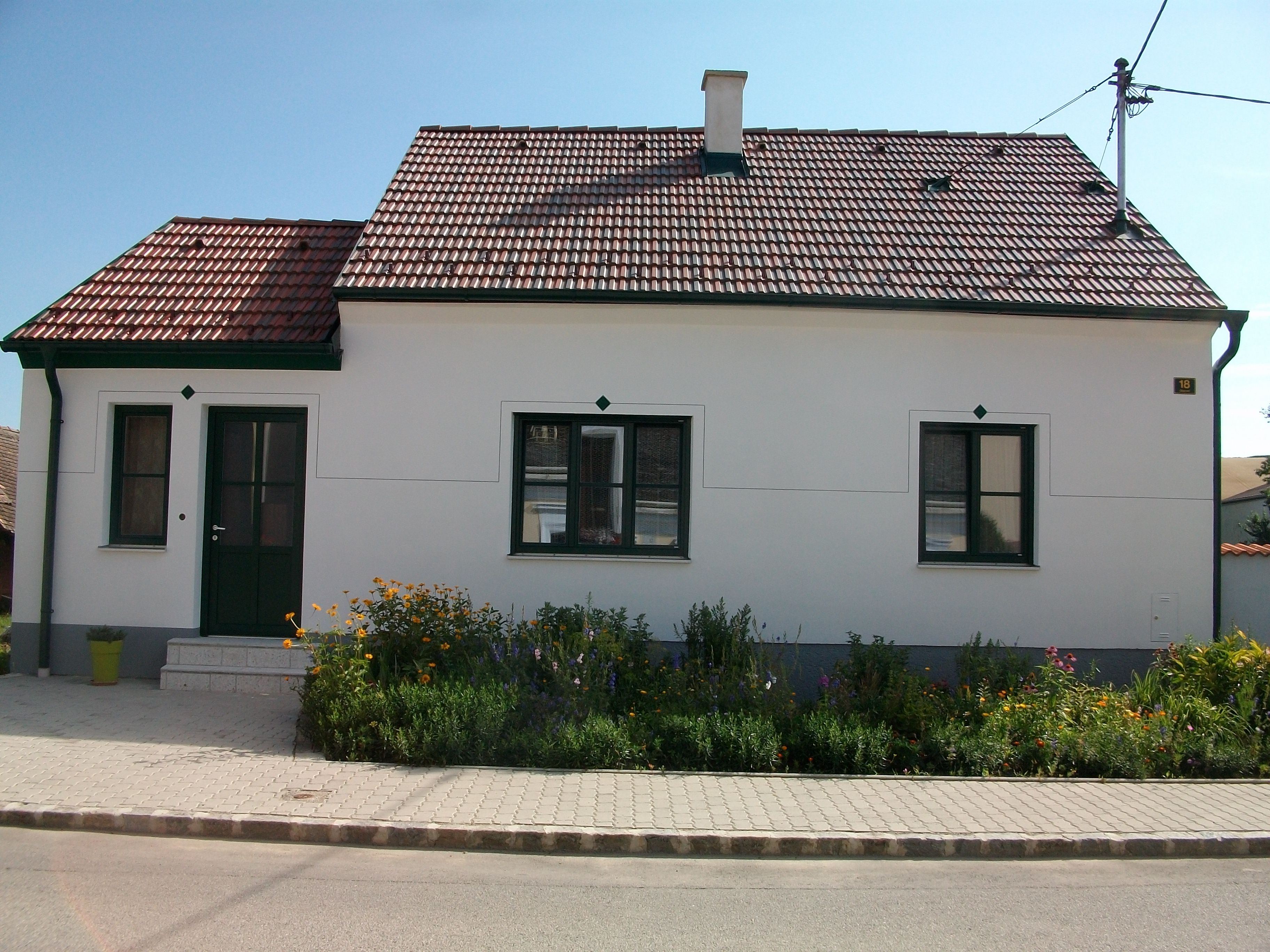 Ein kleines, weißes Haus mit rotem Ziegeldach und grünem Türrahmen, umgeben von einem gepflegten Garten mit Blumen.