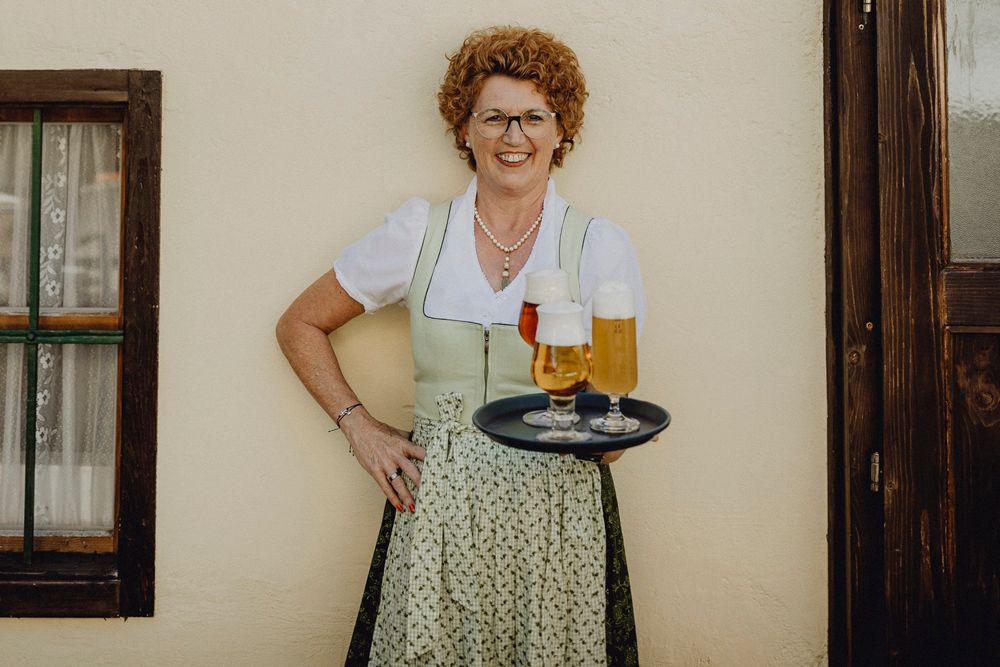 Frau in traditioneller Kleidung mit Tablett voller Biergläser vor einer Wand.