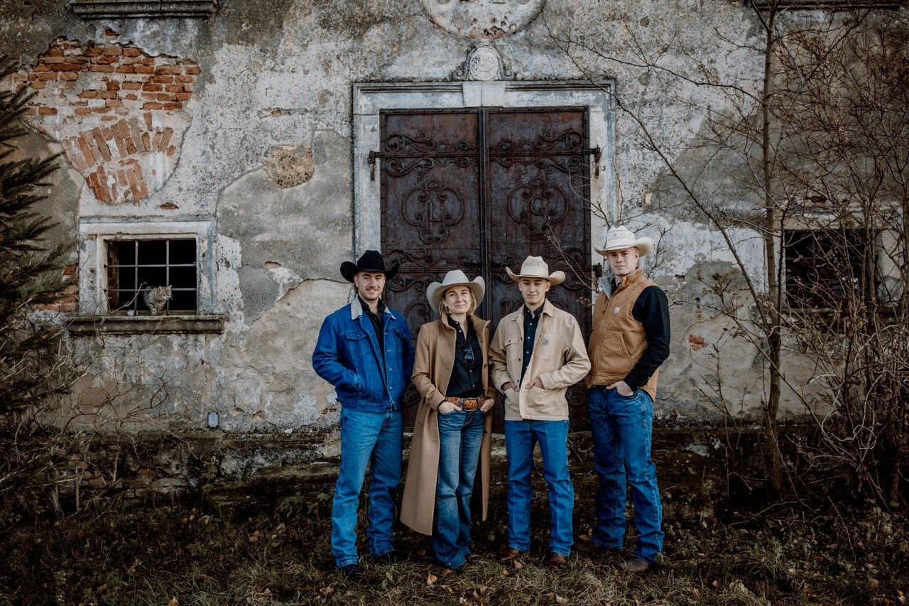 Vier Personen in Cowboykleidung stehen vor einer alten, verfallenen Mauer mit einer großen Metalltür.