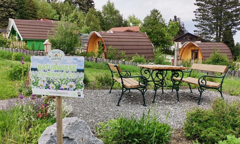 Ein Schild mit der Aufschrift 'Naturwiese für Biene & Co' steht vor einer Wiese mit Bänken und Tischen, umgeben von kleinen, hölzernen Hütten.