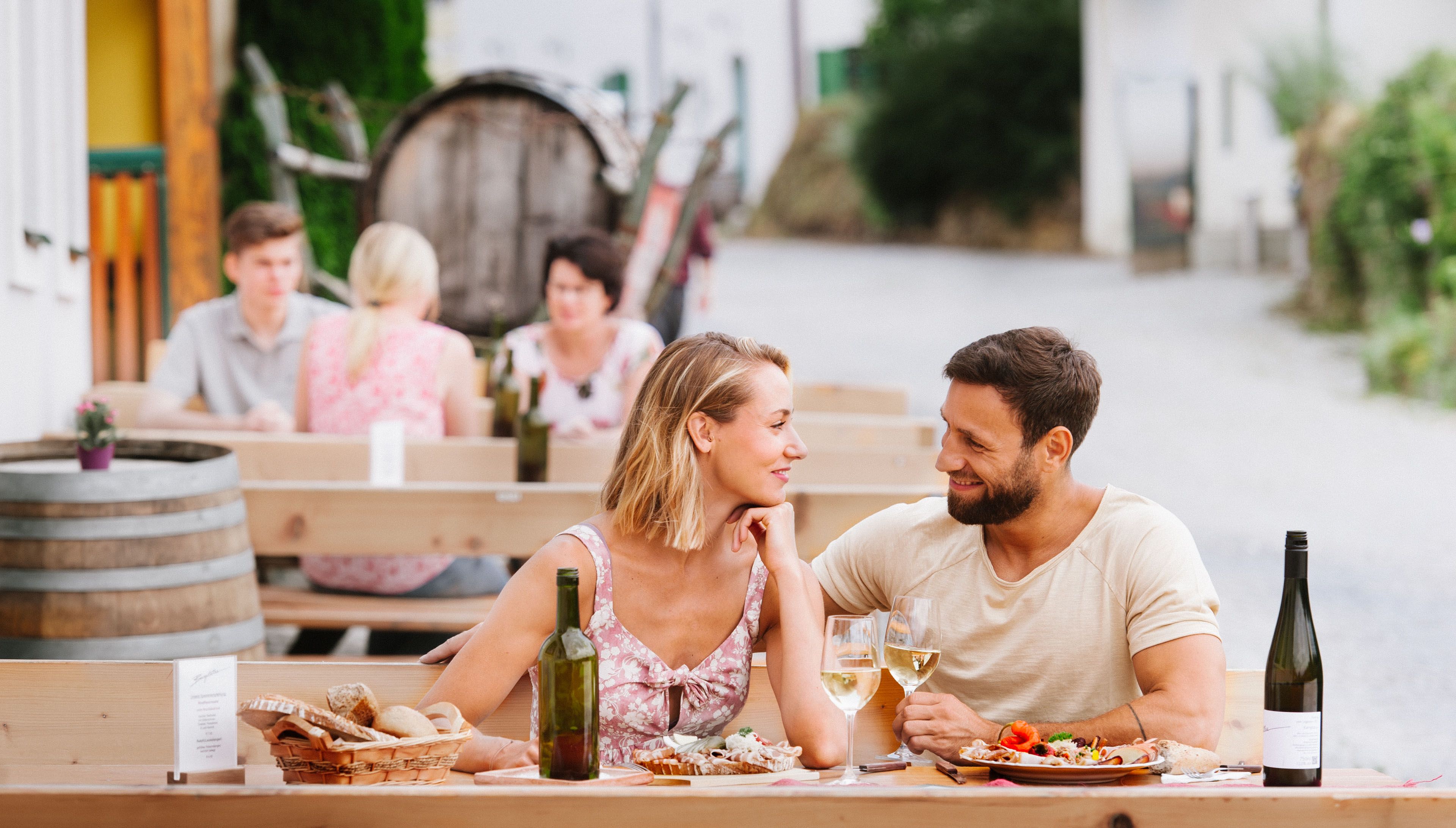 Ein Paar sitzt an einem Tisch im Freien, genießt Wein und Essen. Im Hintergrund sind weitere Gäste und ein Weinfass zu sehen.