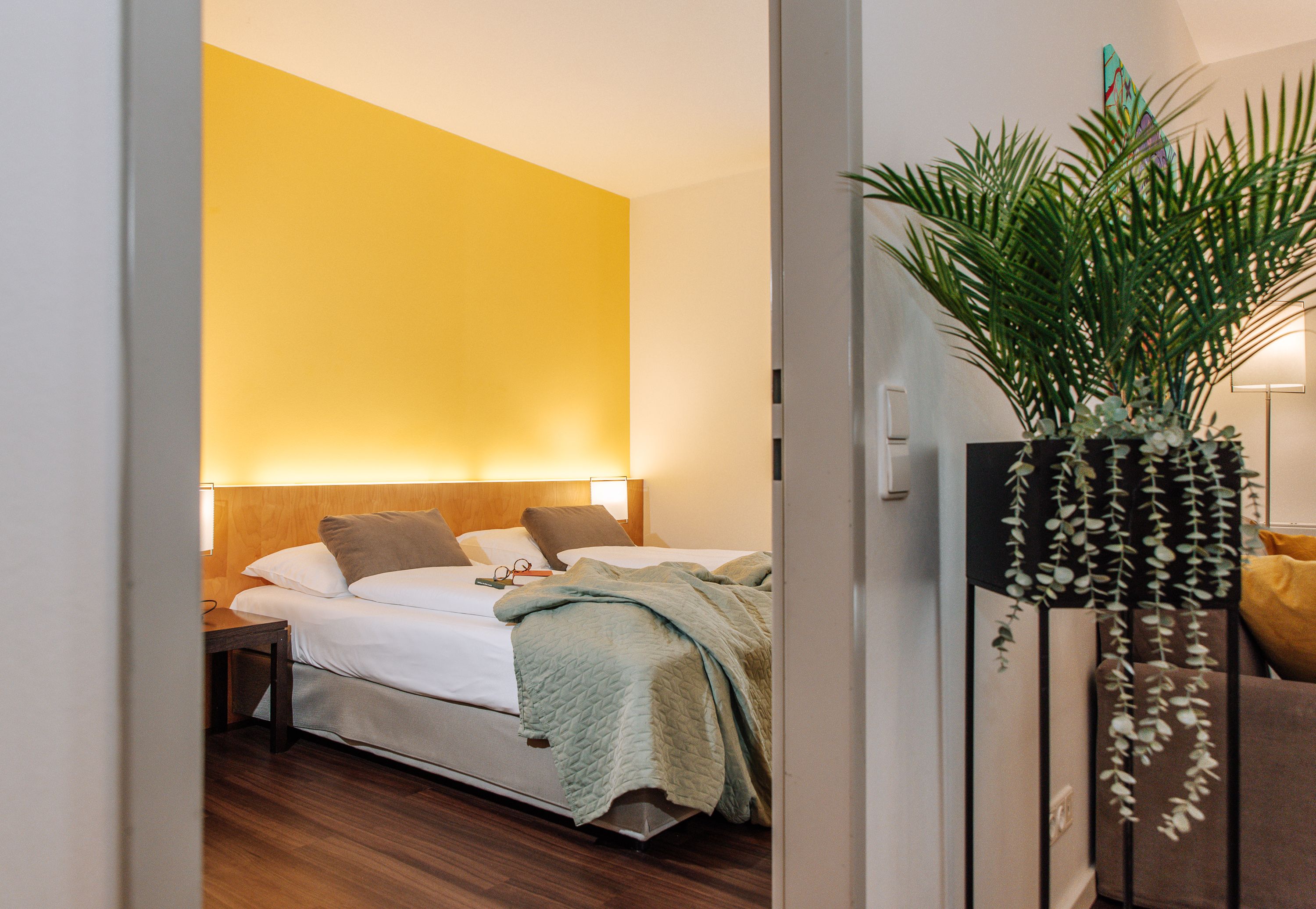 Hotelzimmer mit gelber Wand, Doppelbett und Pflanze im Vordergrund.