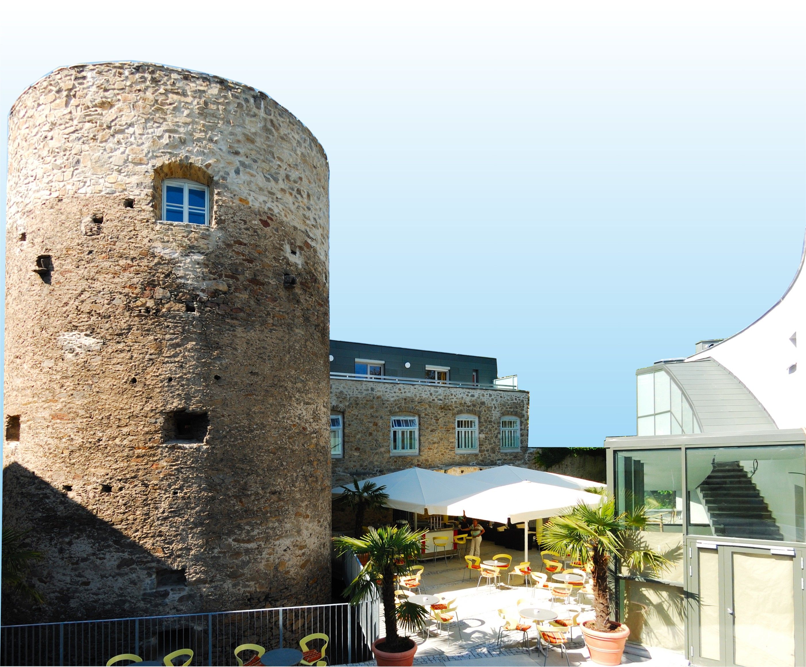 Historischer Turm neben modernem Gebäude mit Terrasse und Sonnenschirmen.