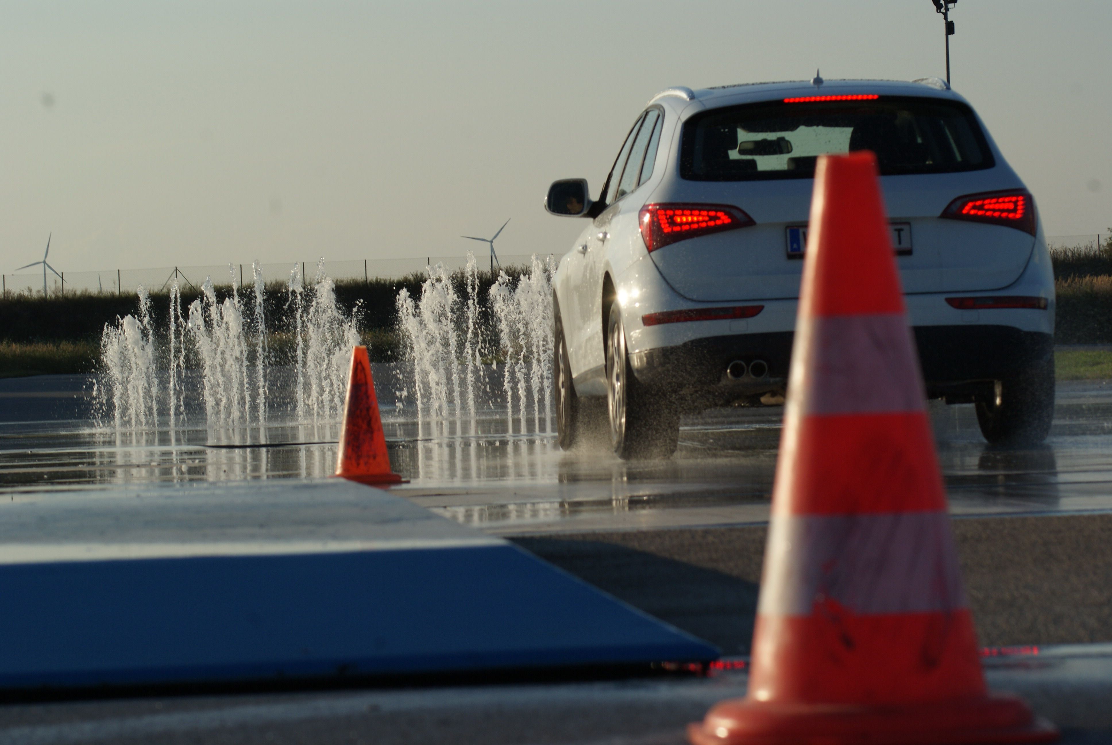 Ein Auto fährt auf einer nassen Fahrbahn mit Wasserfontänen und Verkehrskegeln.