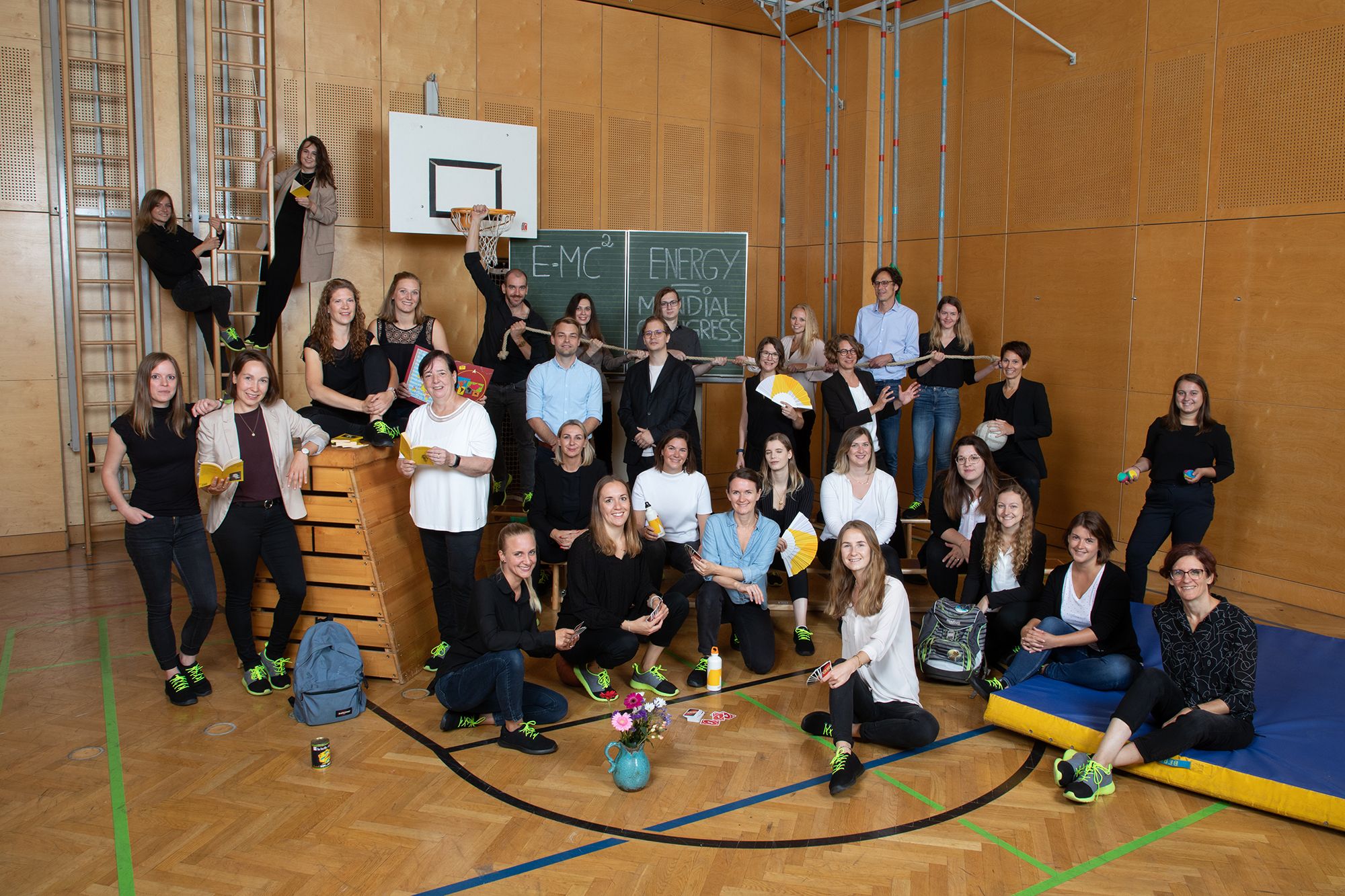 Gruppe von Menschen in einer Turnhalle mit Sportgeräten und Tafel im Hintergrund.