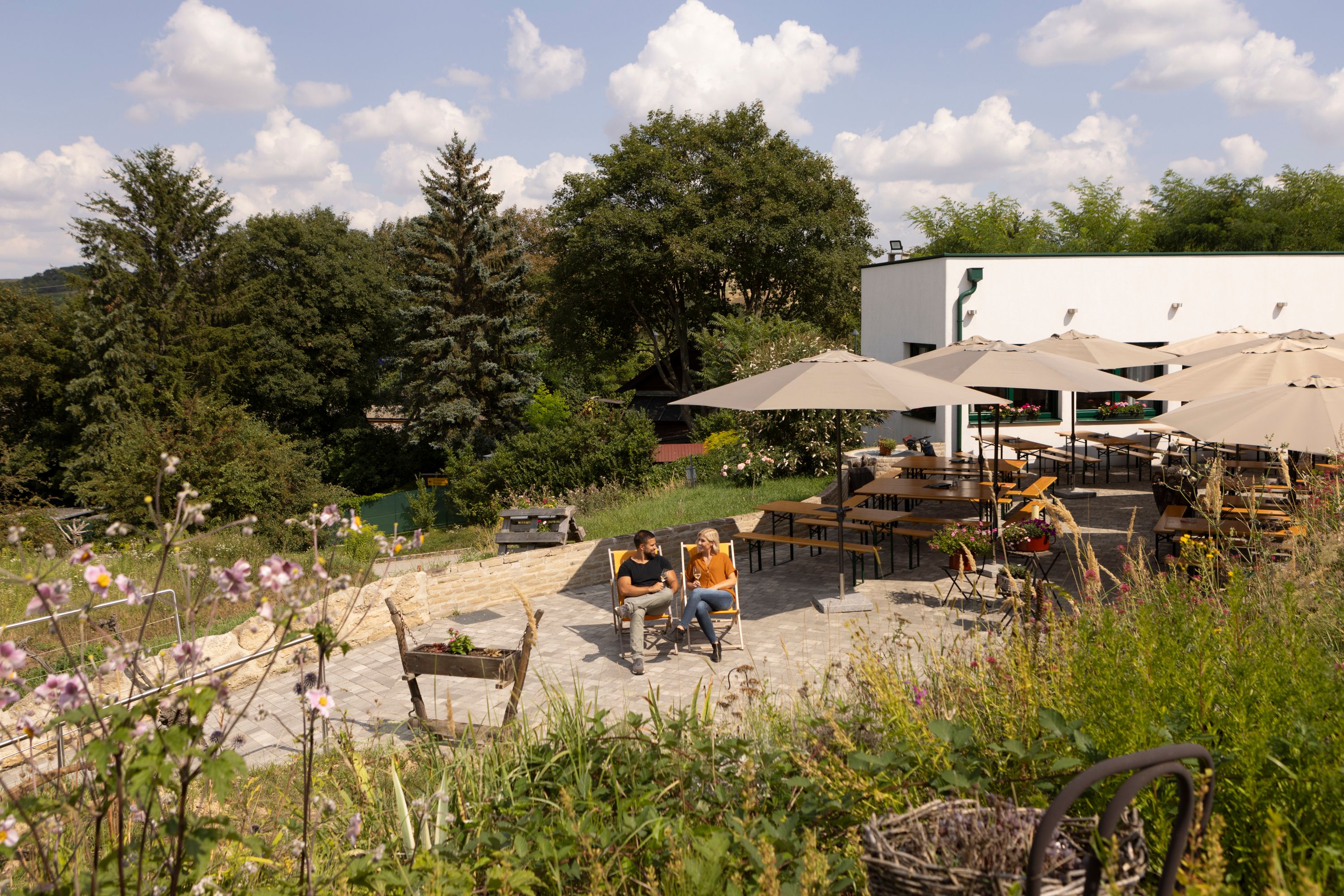 Zwei Personen sitzen auf Liegestühlen in einem Garten mit Sonnenschirmen und Tischen, umgeben von Bäumen und Blumen.