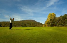 Golfer schlägt Ball auf einem grünen Golfplatz mit bewaldeten Hügeln im Hintergrund.