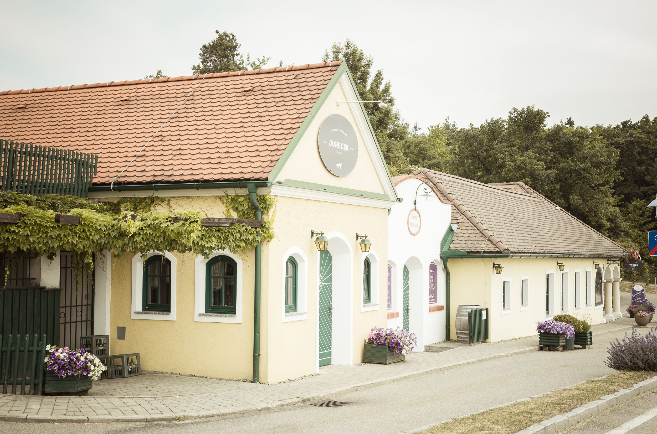Außenansicht eines traditionellen Heurigen mit gelber Fassade und roten Dachziegeln, umgeben von Pflanzen.
