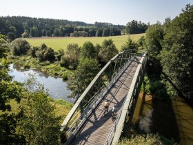 Die sanfte Brücke schwingt über den glitzernden Fluss, umgeben von üppigem Grün und der Ruhe der Natur. Radfahrer genießen die frische Luft und die malerische Landschaft, während die Sonne sanft auf die Wiesen scheint.