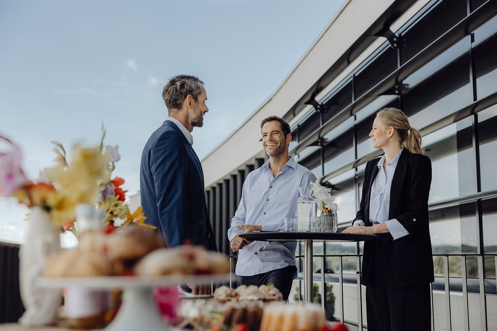 Drei Personen unterhalten sich auf einer Terrasse neben einem modernen Gebäude.
