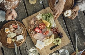 Ein rustikaler Holztisch mit einer Jause aus Brot, K&auml;se, Wurst, Gem&uuml;se und Wein.