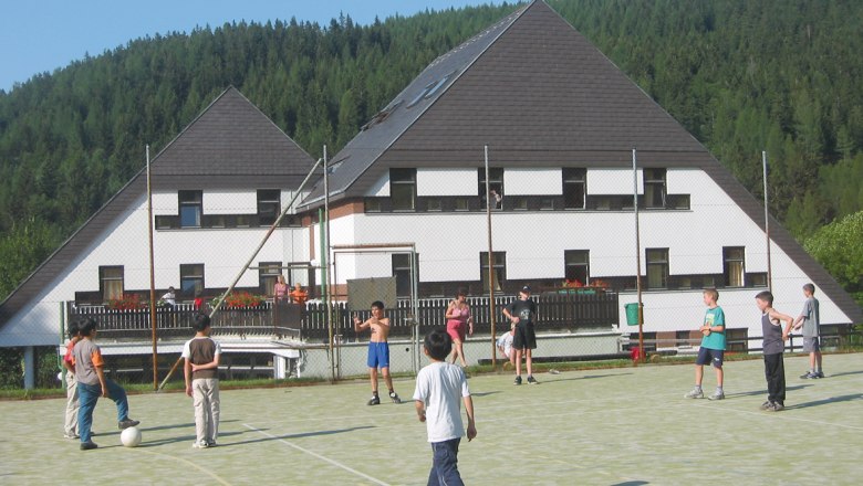 Junges Hotel Annaberg, © zVg Gemeinde Annaberg Kinder spielen Fußball auf einem Sportplatz vor einem Hotel in einer bewaldeten Gegend.