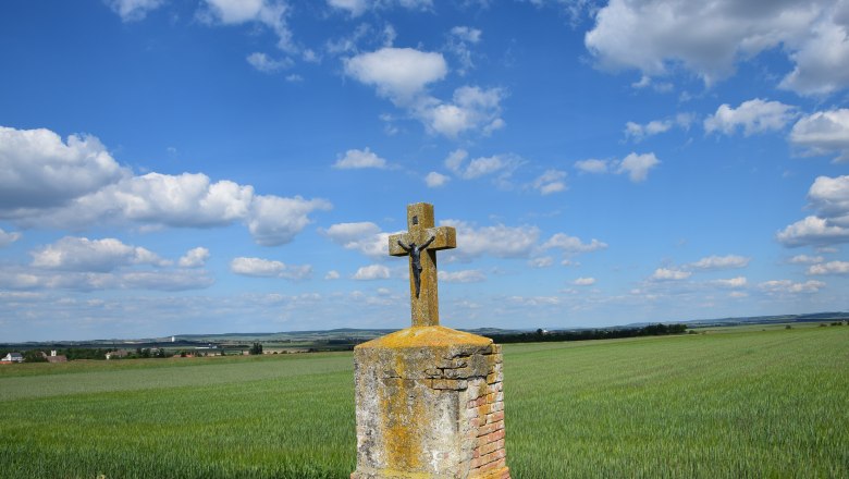 Ein steinernes Kreuz mit Christusfigur steht auf einem Feld unter blauem Himmel mit Wolken.