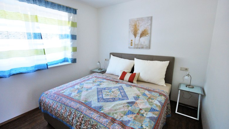 Ein Schlafzimmer mit einem Doppelbett, buntem Bett&uuml;berwurf, zwei Nachttischen und einem Fenster mit gestreiften Vorh&auml;ngen.