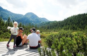 Familie sitzt auf Holzplattform im Hochmoor Leckermoos, umgeben von Bergen und Wald.