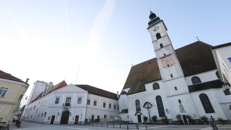 Pfarrkirche mit Uhrturm und angrenzenden Gebäuden auf einem leeren Platz.
