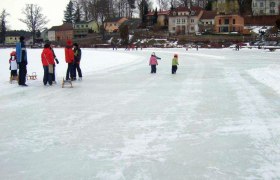 Menschen eislaufen auf einem zugefrorenen See in Allentsteig, umgeben von H&auml;usern und B&auml;umen.