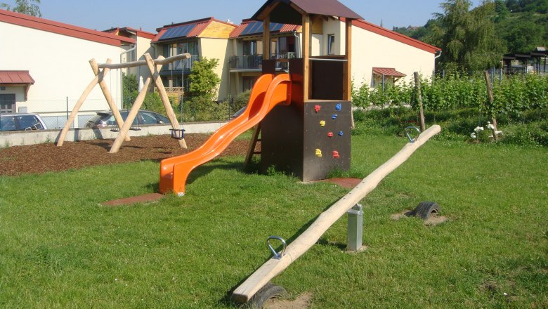 Ein Kinderspielplatz mit Rutsche, Schaukel und Wippe auf einer Wiese vor Wohnhäusern.