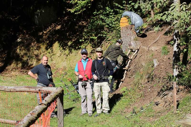 Drei Männer mit Bögen im Wald, ein Mann klettert eine Leiter hoch.