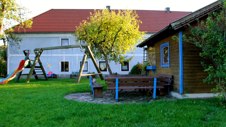 Ein Spielplatz mit Schaukel, Rutsche und Sitzb&auml;nken vor einem Haus mit rotem Dach.