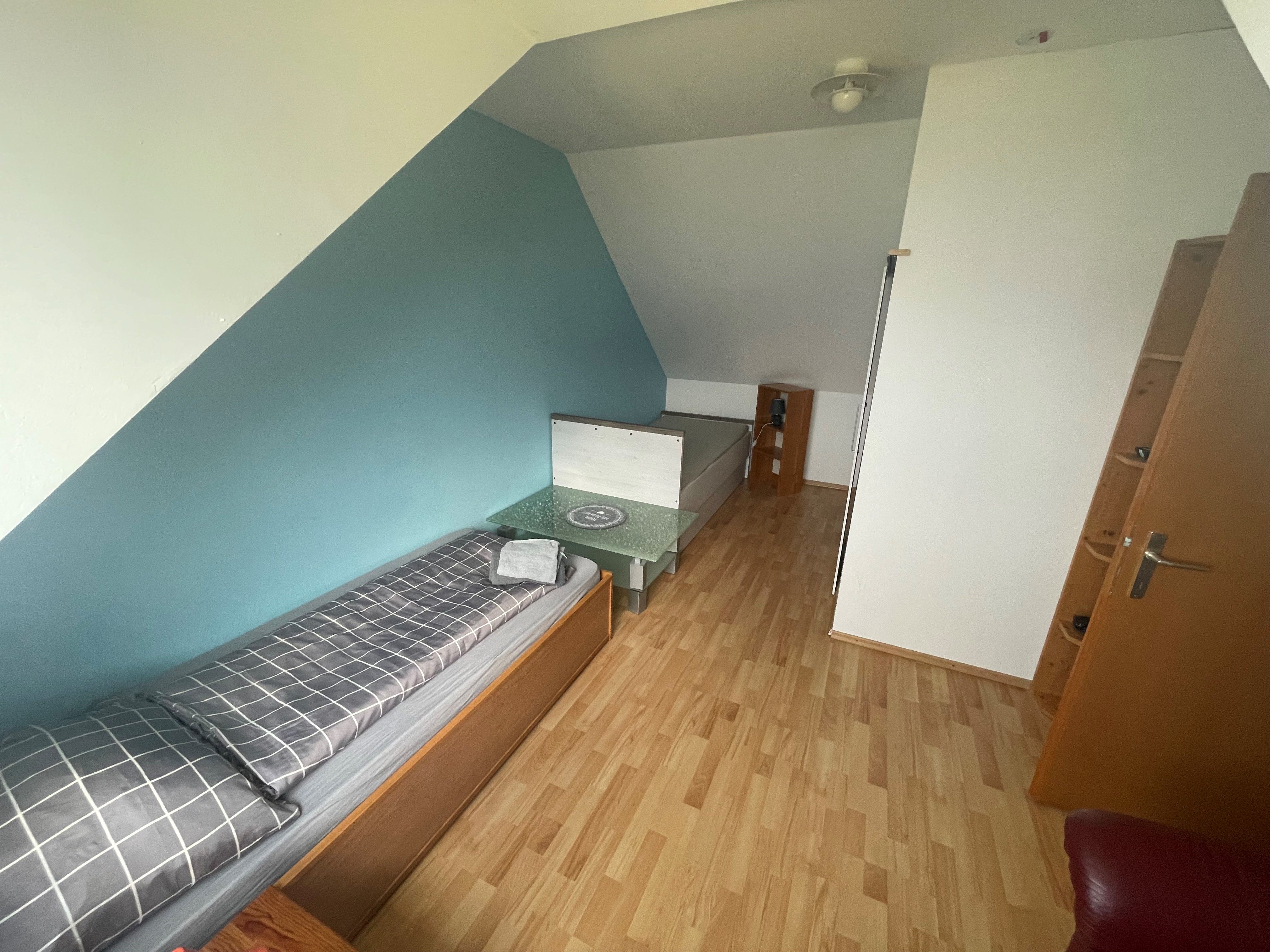 Ein Zimmer mit einem Einzelbett, einem kleinen Tisch und einem Regal. Die Wände sind blau und weiß gestrichen.