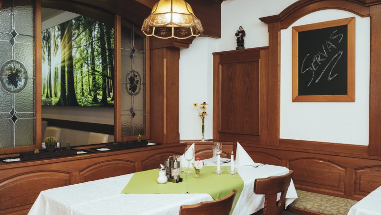 Gem&uuml;tlicher Gastraum mit Holzm&ouml;beln, Tisch mit gr&uuml;ner Tischdecke, Kerzen und Dekoration. Fenster mit Waldmotiv und Tafel mit Schriftzug 'Servas'.