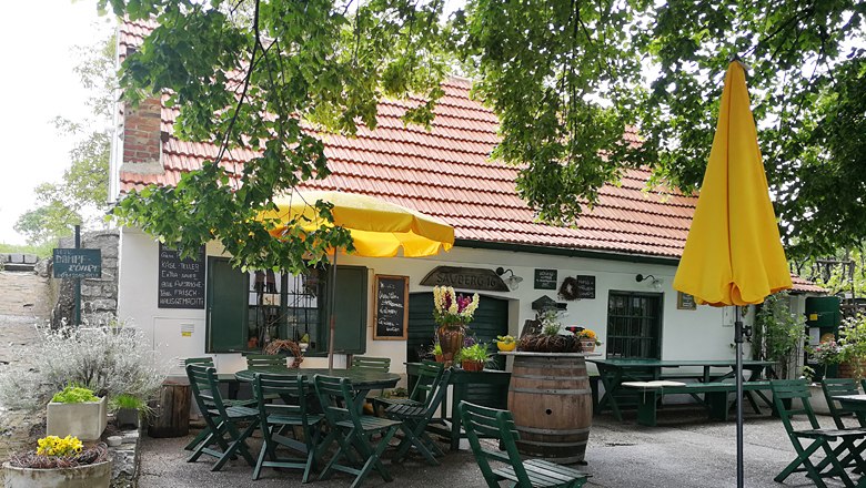 Gem&uuml;tlicher Gastgarten mit gelbem Sonnenschirm und Holztischen vor einem Geb&auml;ude mit rotem Ziegeldach.