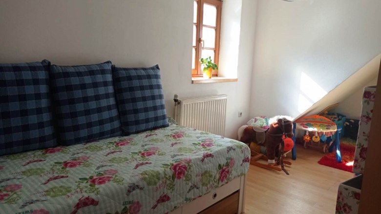 Kinderzimmer mit ausziehbarem Bett, karierten Kissen, Spielzeugpferd und Spieltisch. Helles Fenster mit Pflanze.