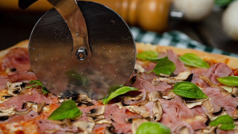 Nahaufnahme einer Pizza mit Schinken, Pilzen und Basilikum, die mit einem Pizzaschneider geschnitten wird.