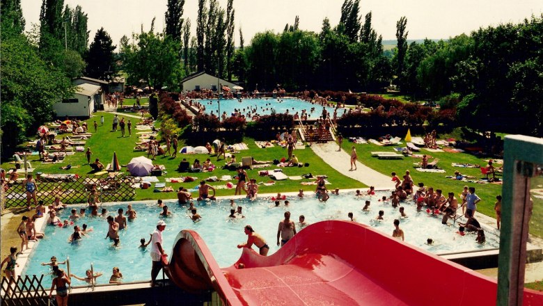 Freibad mit vielen Menschen im Wasser und auf der Liegewiese, rote Rutsche im Vordergrund.