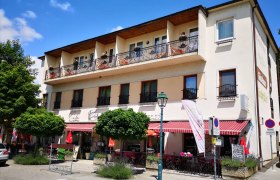 Außenansicht eines Cafés mit Balkon und Markise, umgeben von Bäumen und Straßenschildern.