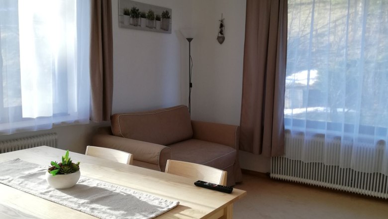 Helles Wohnzimmer mit Esstisch, St&uuml;hlen, Sofa und gro&szlig;en Fenstern mit Vorh&auml;ngen.