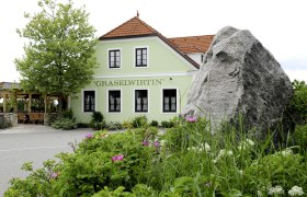Grünes Gebäude mit der Aufschrift 'Graselwirtin', umgeben von Pflanzen und einem großen Felsen im Vordergrund.